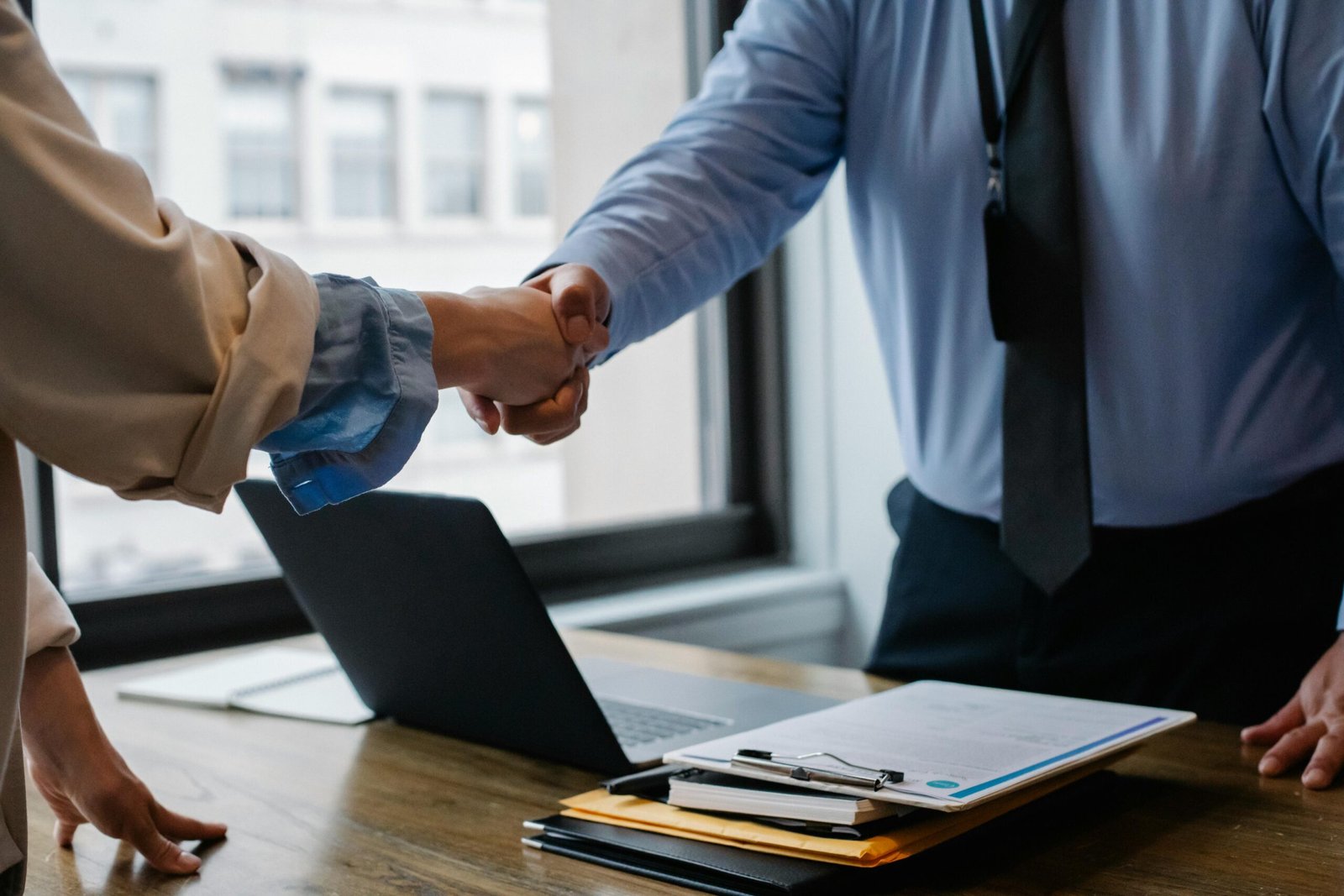 my home Crop unrecognizable coworkers in formal wear standing at table with laptop and documents while greeting each other before meeting